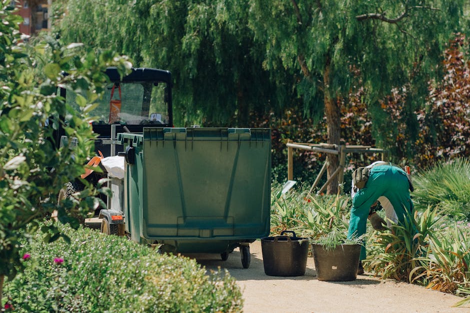 A gardener wearing teal overalls is crouched down, tending to a garden bed filled with ornamental plants and foliage. Nearby, two large black containers hold trimmed plant cuttings or garden waste. An